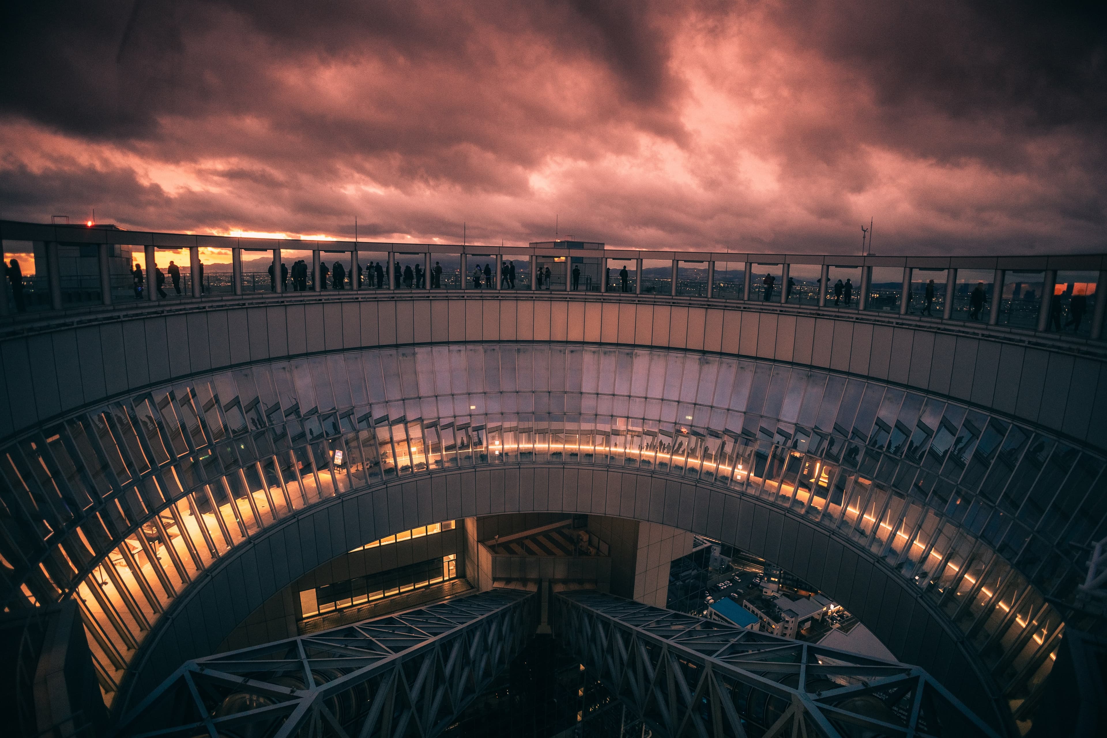 Visitors line the railing of an Osaka observation deck as the sky burns orange through heavy cloud.