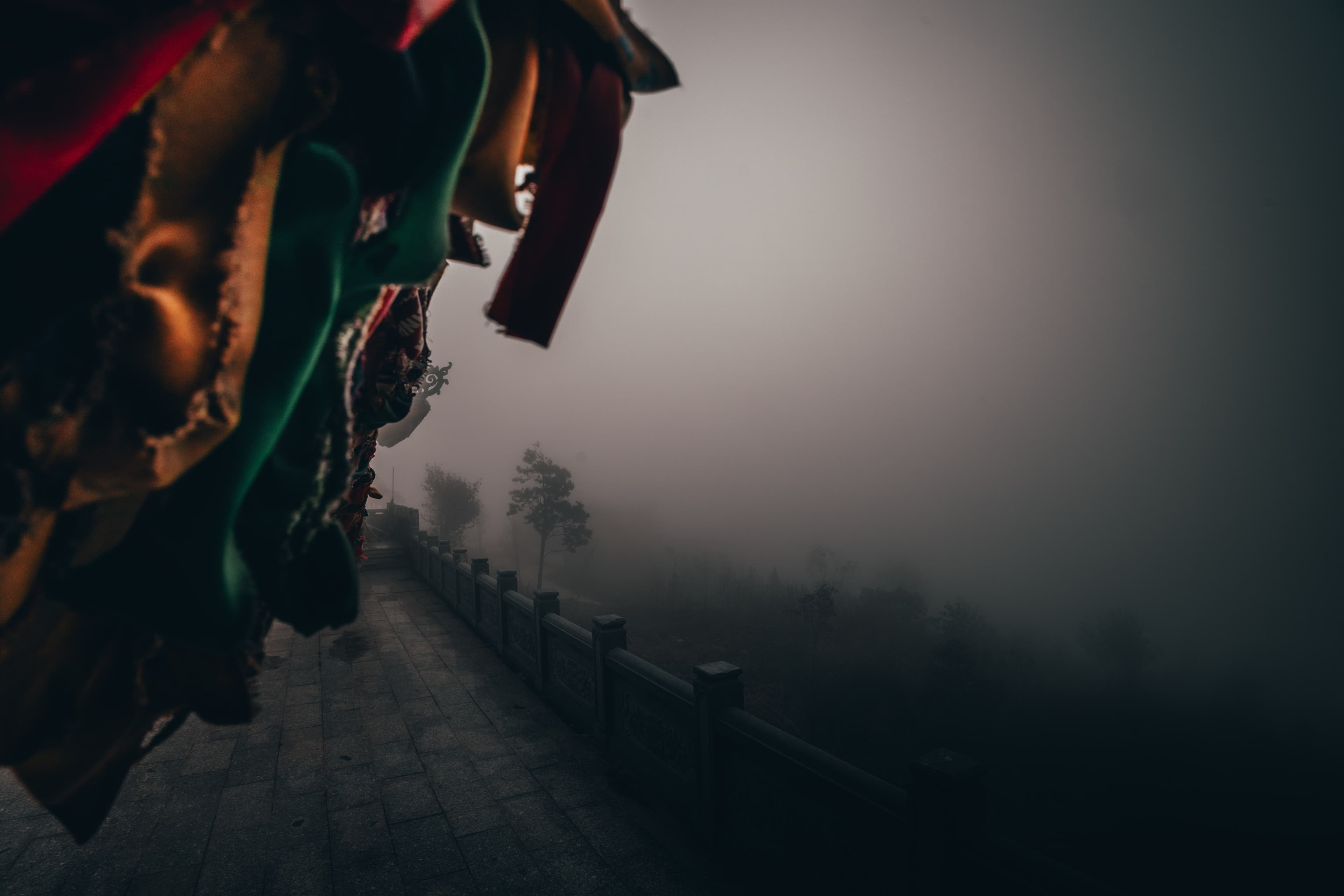 Colorful fabric banners whip in the wind at the edge of a fogged-out overlook, a stone path fading into grey.