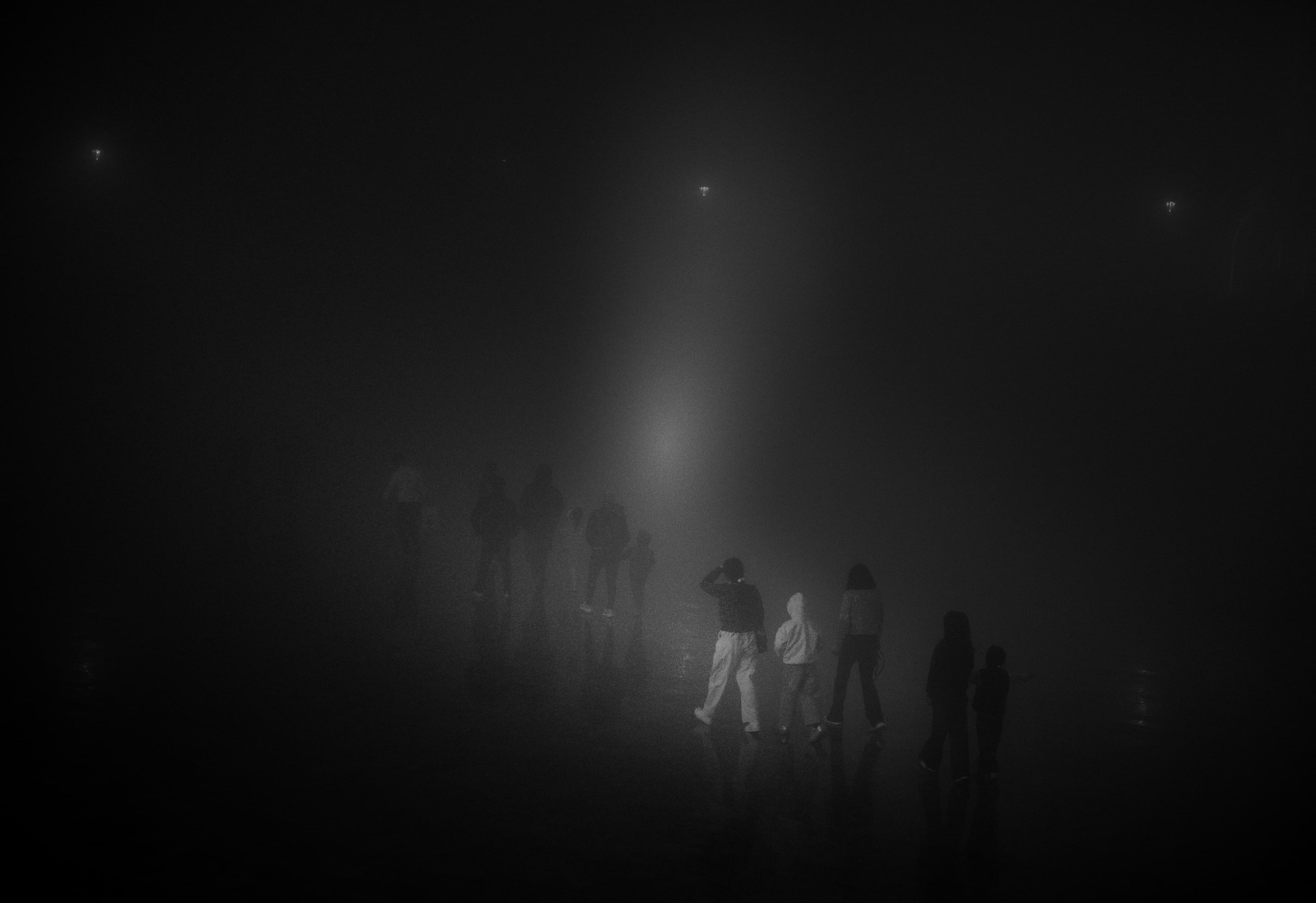 Silhouetted figures gather under a single light in dense fog, barely visible against the dark.