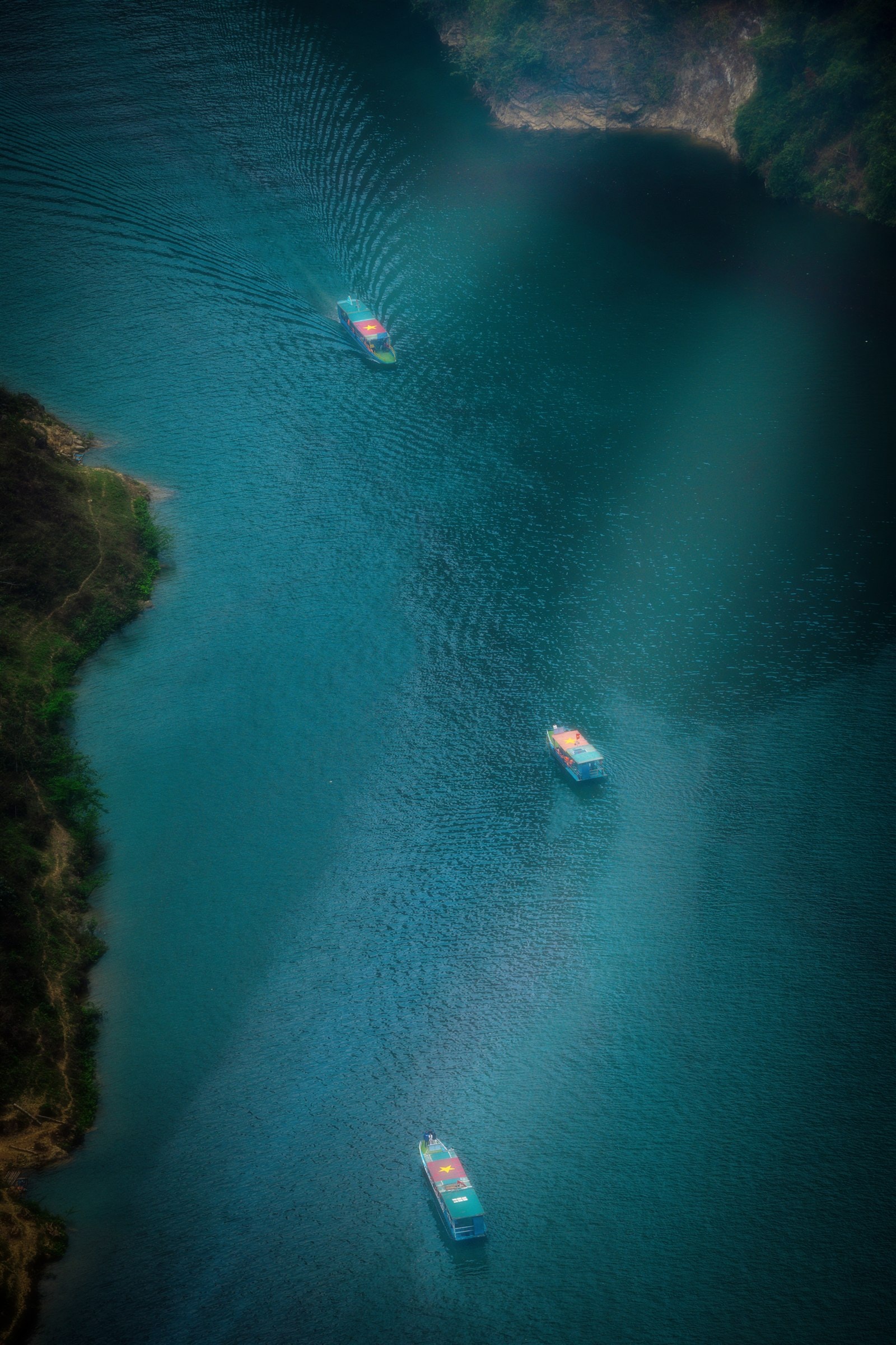 Three boats crossing the turquoise Nho Que river gorge, seen from above with a soft vignette blur.