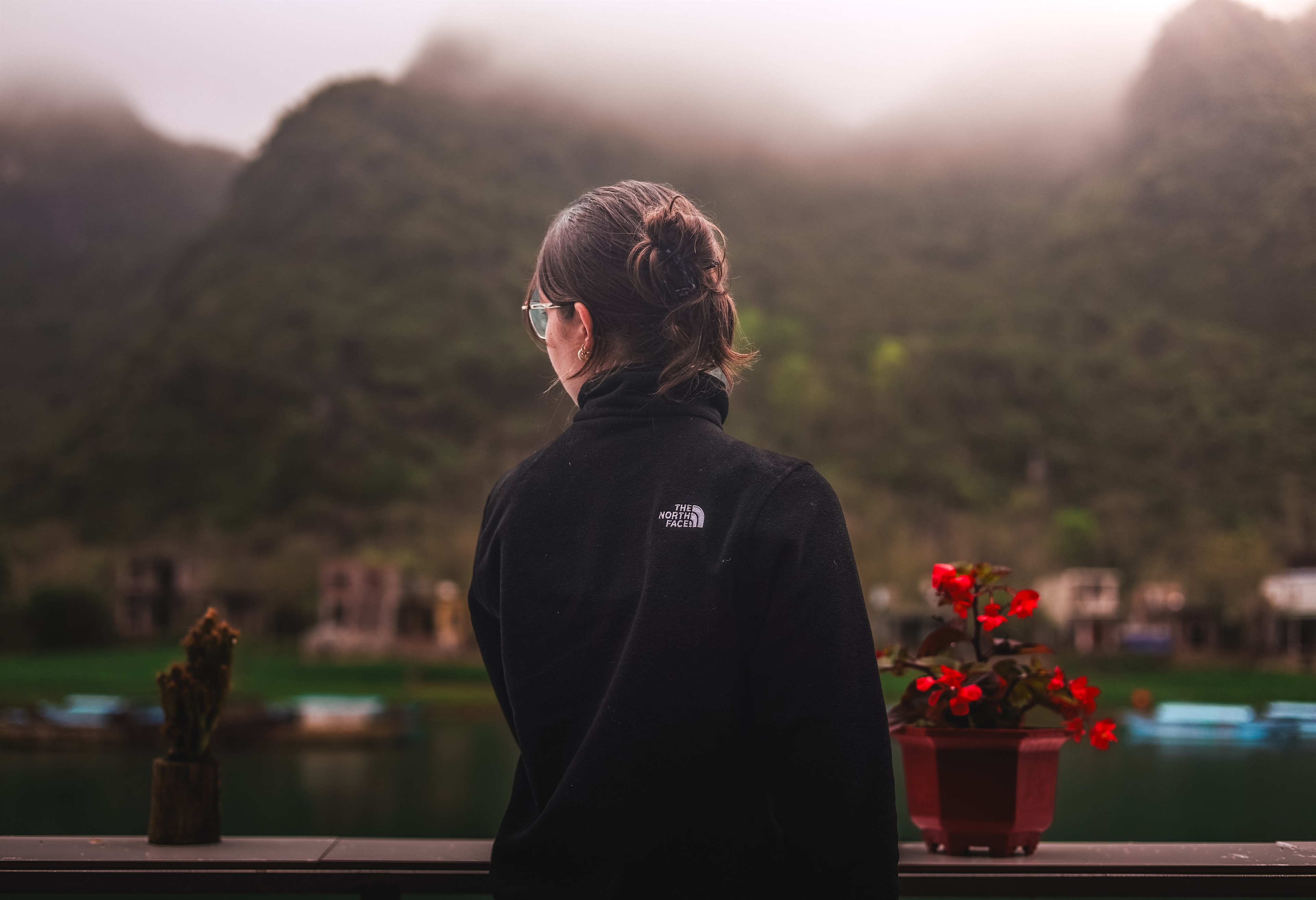 A woman looks out over a misty karst river town from a balcony, red flowers and moored boats below.