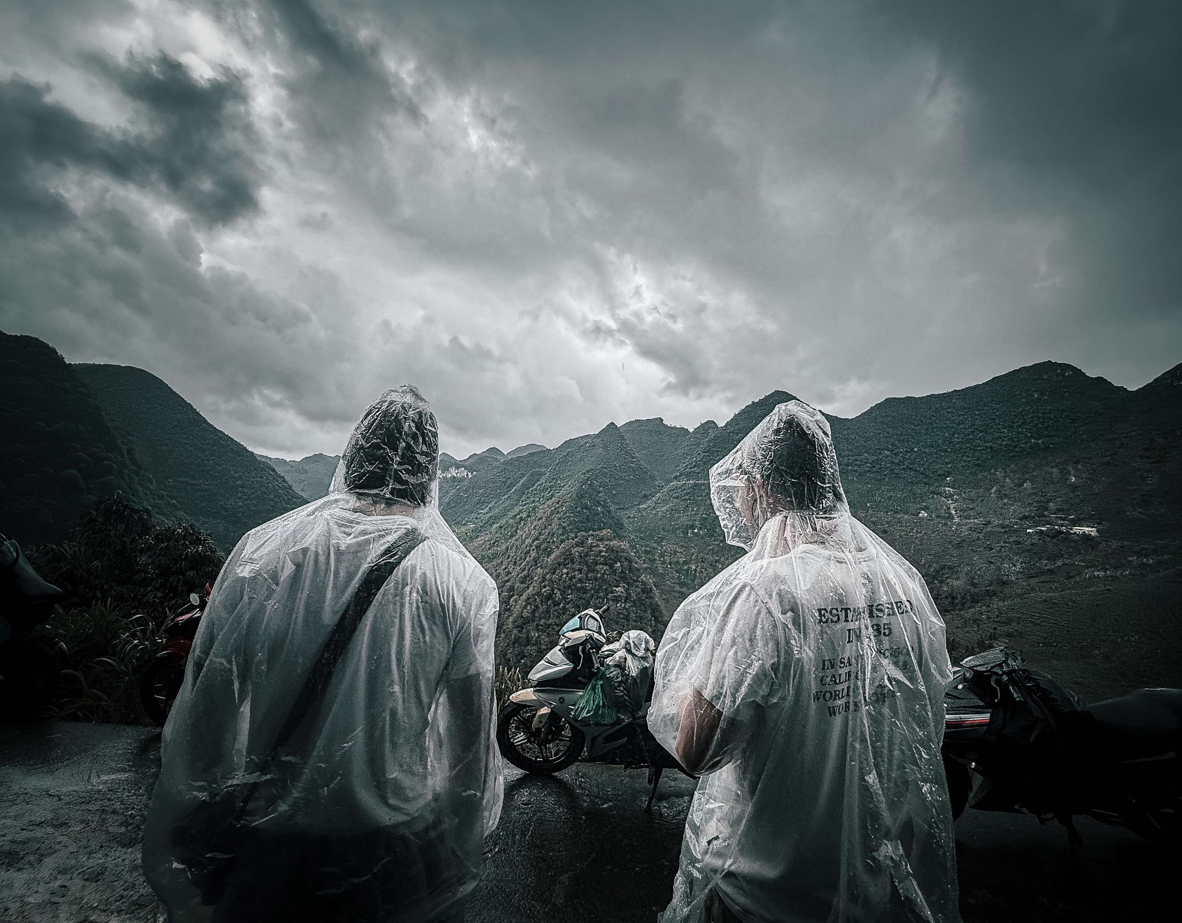 Two riders in clear rain ponchos beside their motorbikes, storm-lit karst peaks behind them.