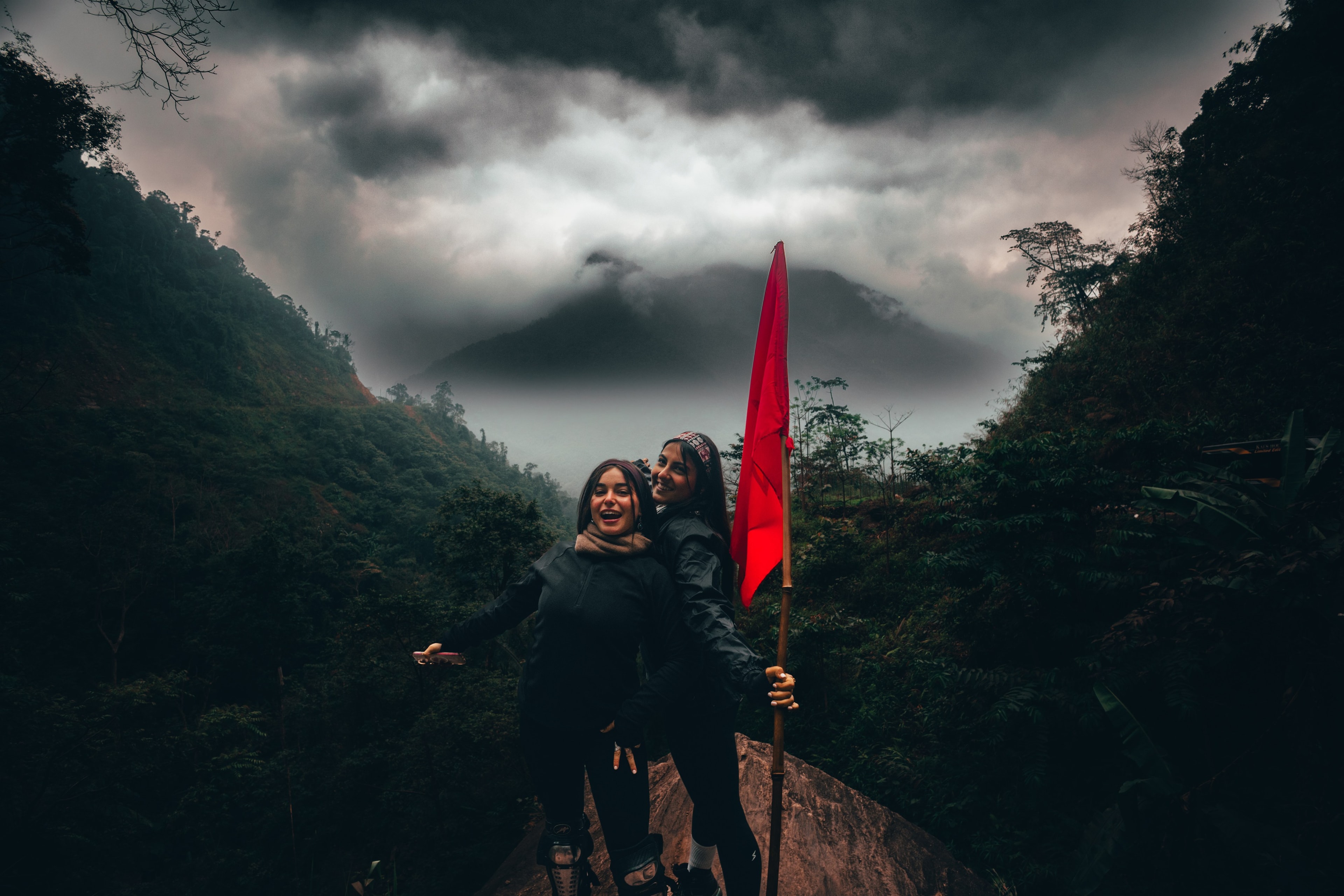Two travelers laughing beside a red flag on a fog-covered ridge, jungle mountains behind them.