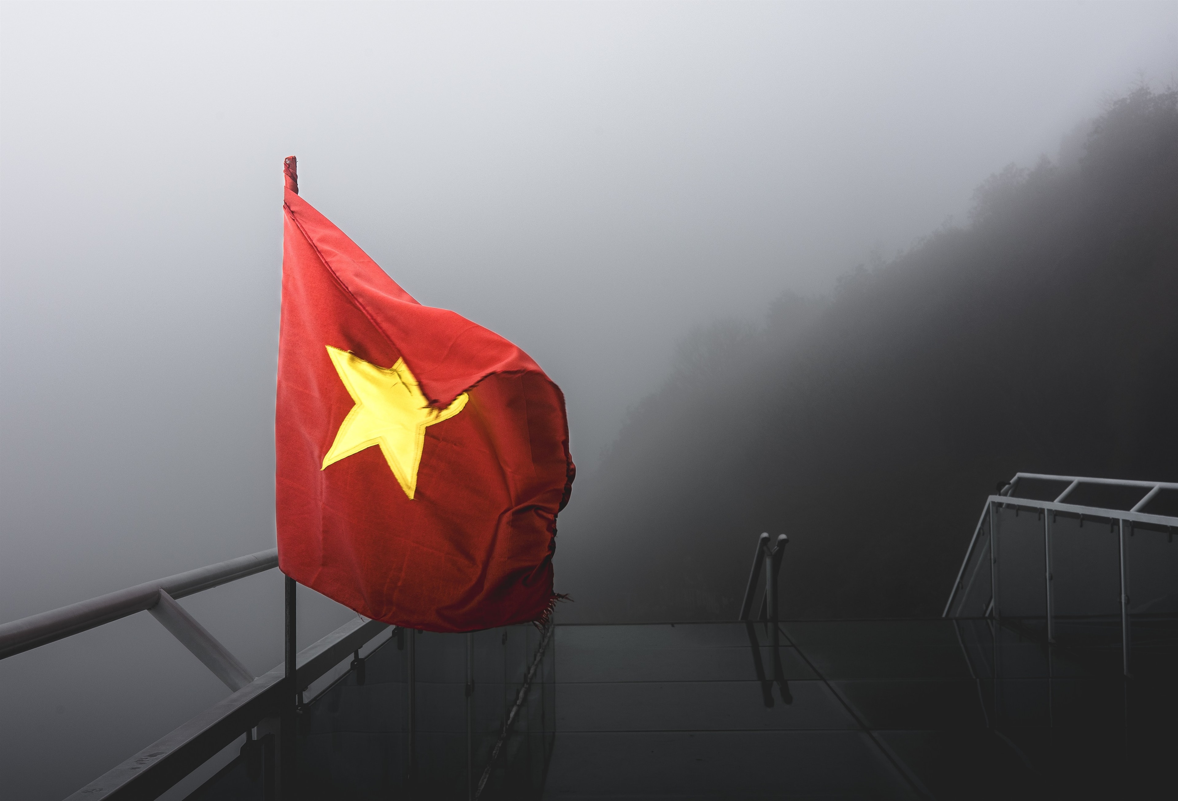 The Vietnamese flag backlit by fog on a mountain overlook, its color the only warmth in a grey landscape.
