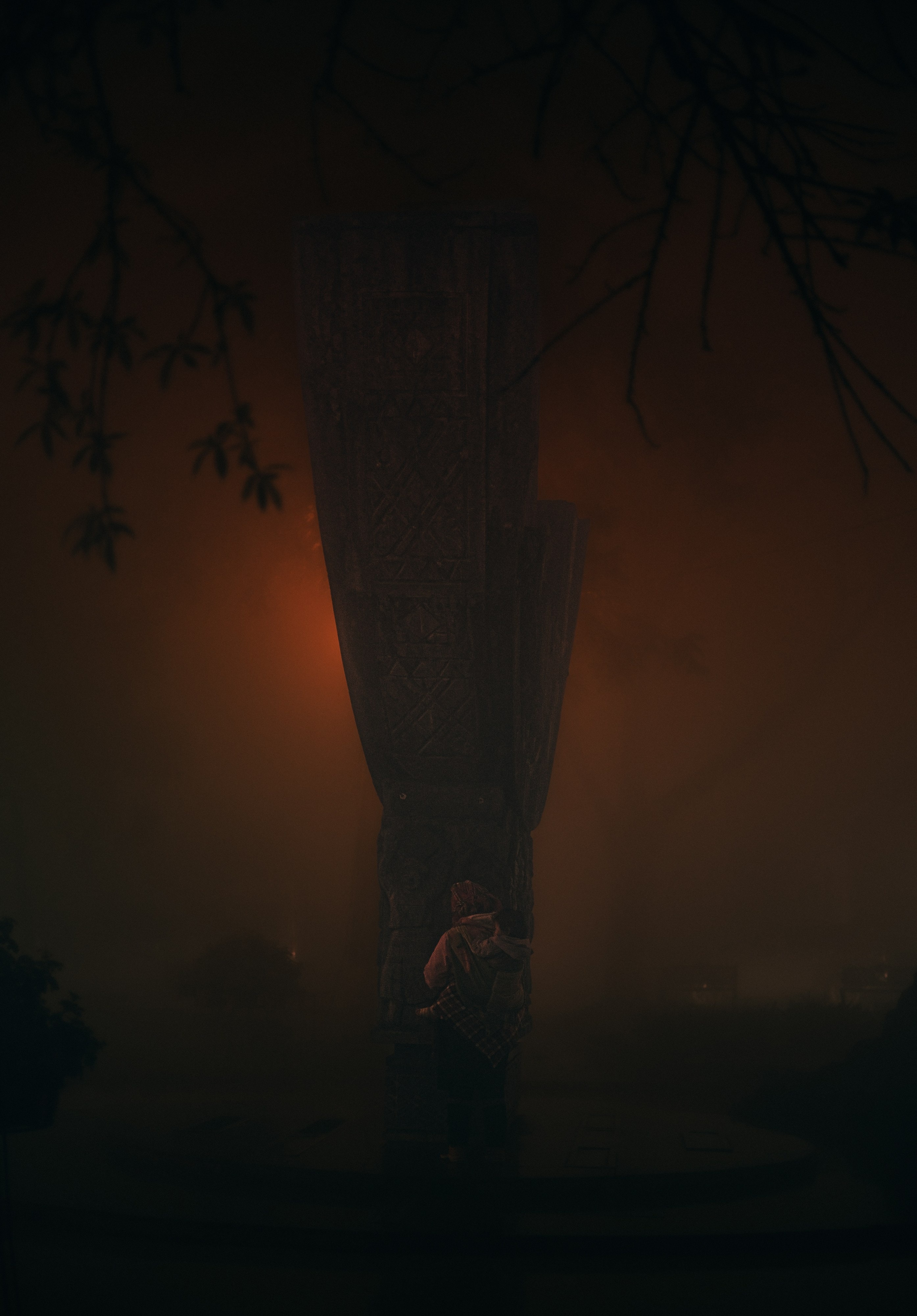 A silhouetted figure against a tall, ornately carved wooden pillar lit in warm orange at night.