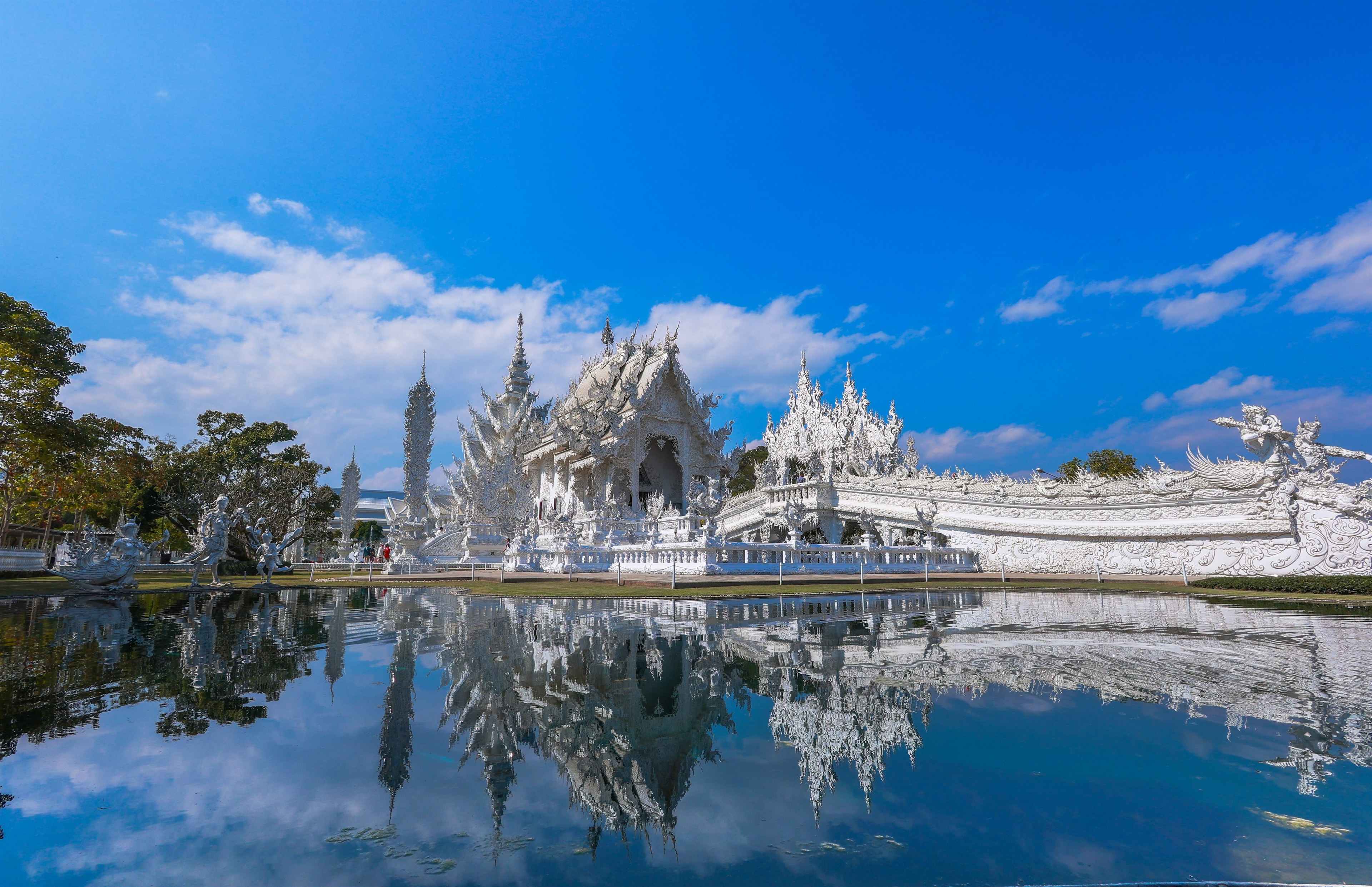 Wat Rong Khun's white spires mirrored in the temple's reflecting pool under a clear sky.