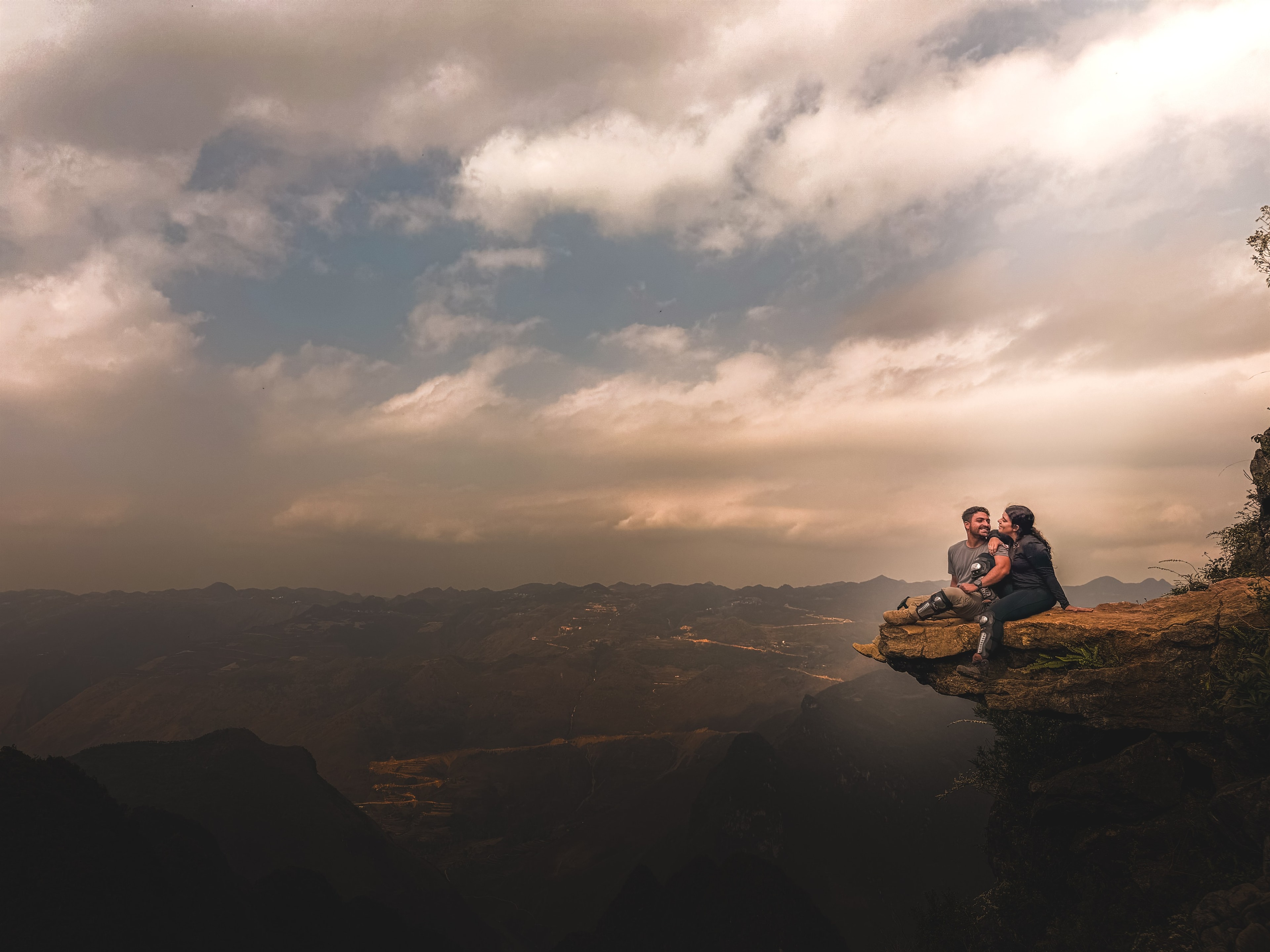 Two travelers sit on a rock ledge above the Ha Giang valley, watching the light change as cloud moves through below.